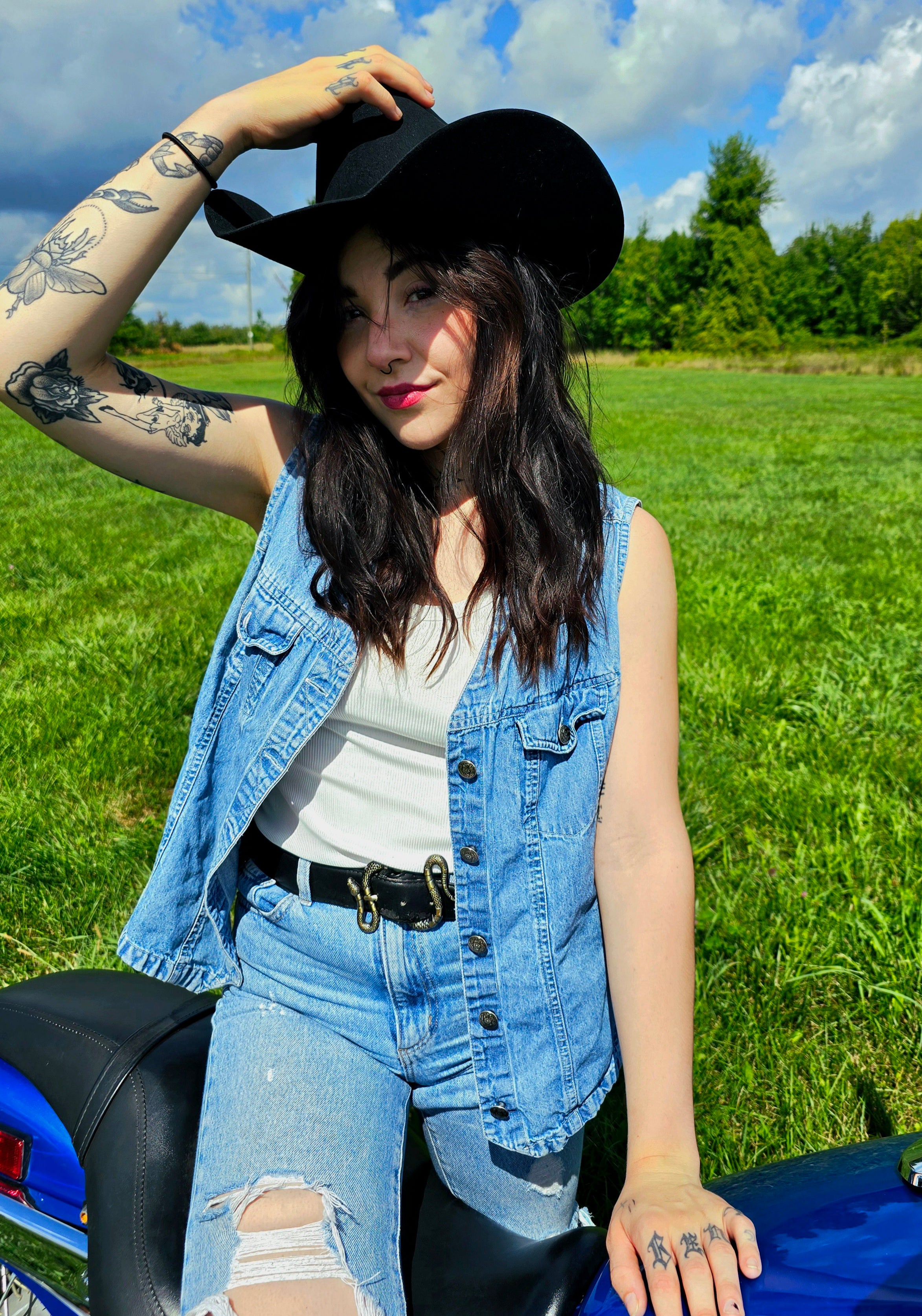Woman wearing a black hat and denim vest sitting on a motorcycle in a grassy field.