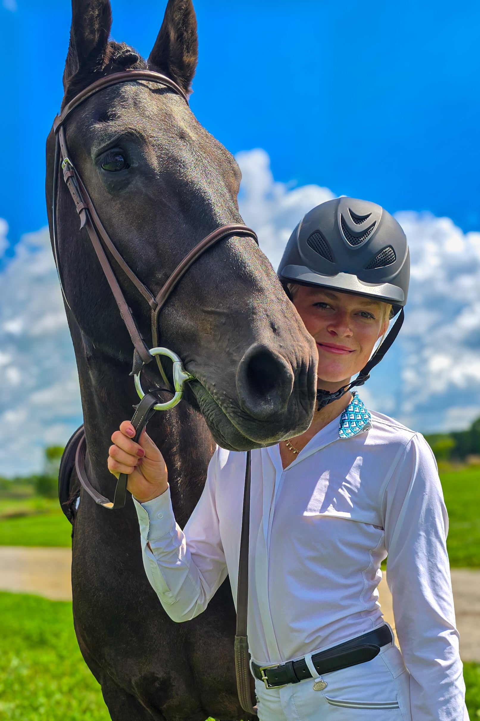 Woman in equestrian attire standing next to a horse with a blue sky background