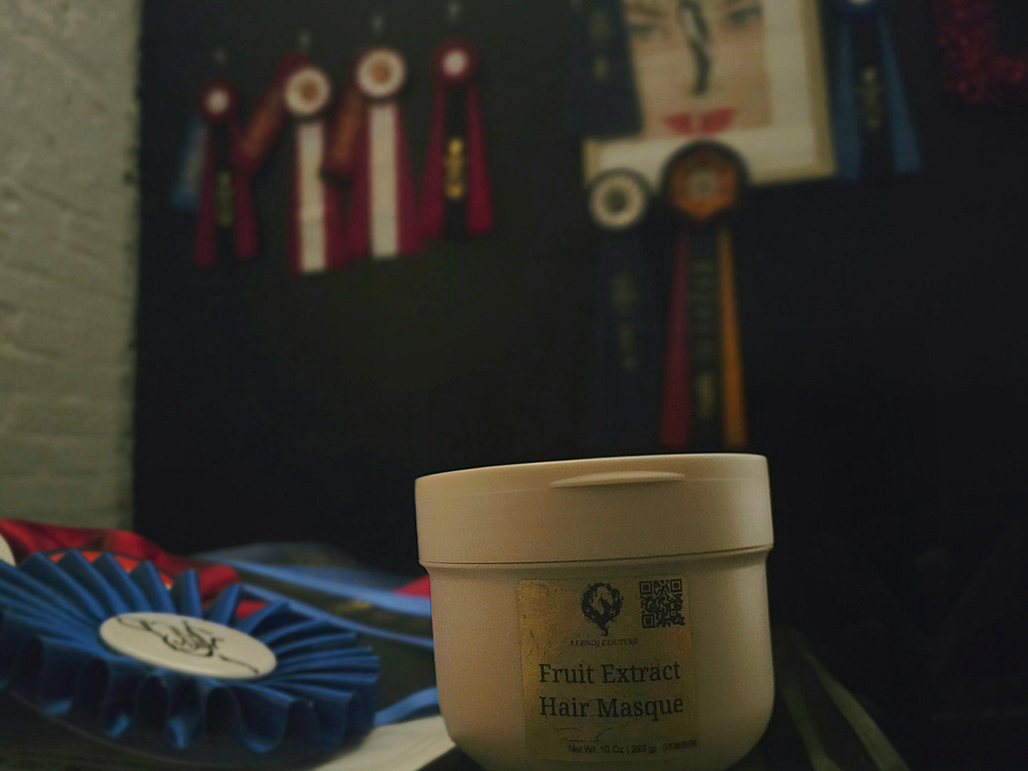 A white plastic jar of Hair Masque with a gold label reading 'Fruit Extract Hair Masque' in the foreground, set against a dark background with blurred ribbons and a picture frame.