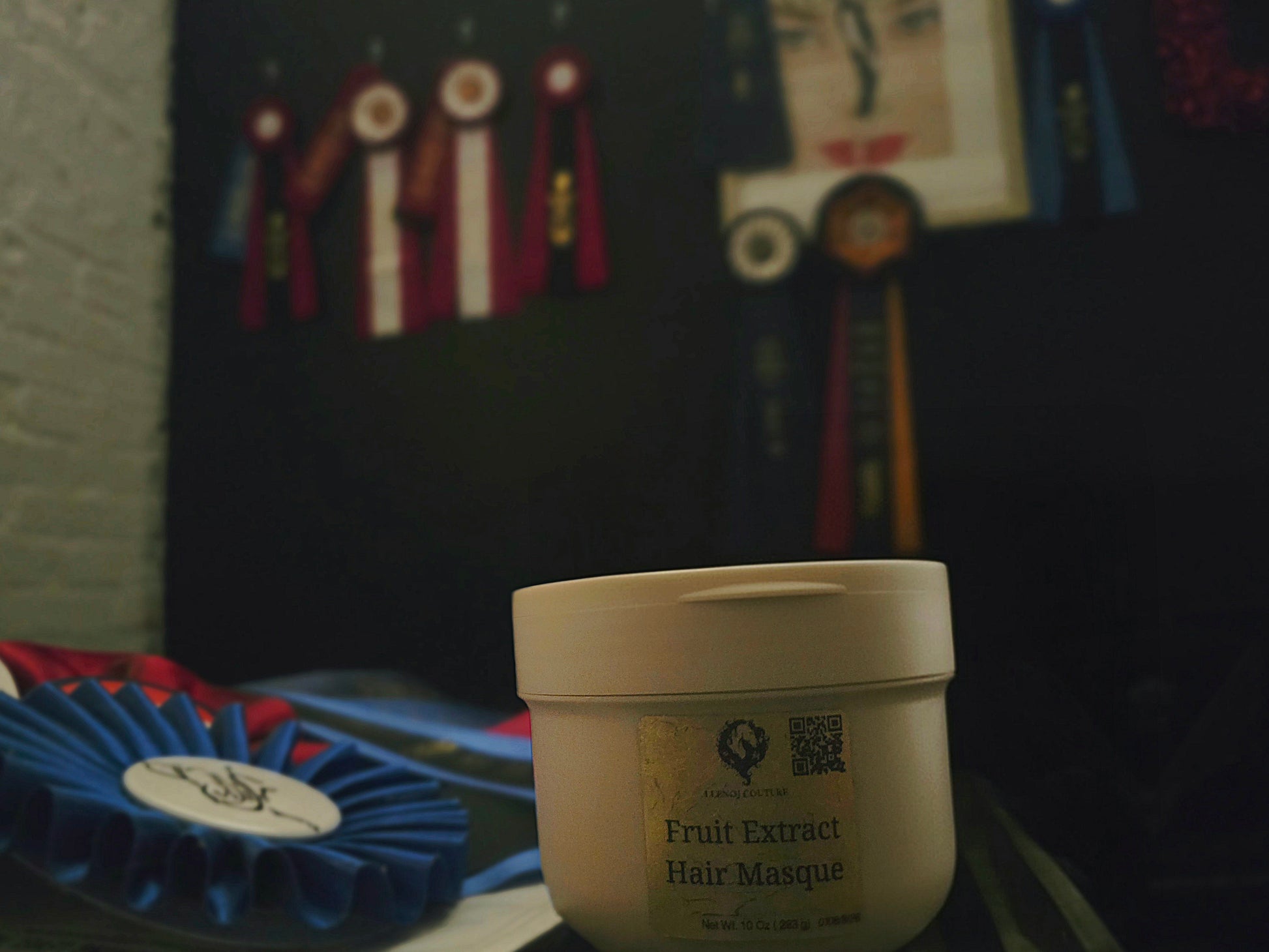 A white plastic jar of Hair Masque with a gold label reading 'Fruit Extract Hair Masque' in the foreground, set against a dark background with blurred ribbons and a picture frame.